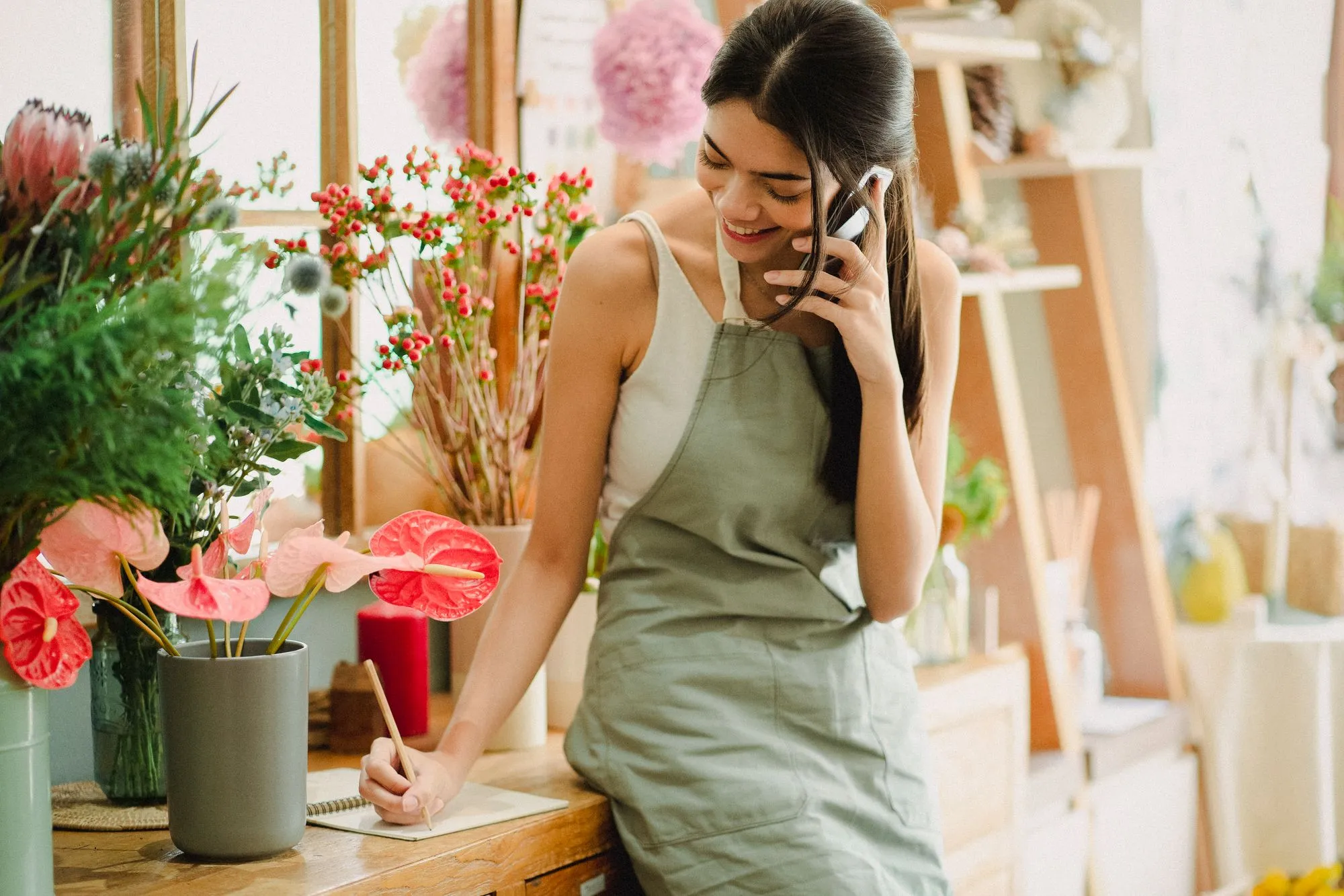 A florist smiling on the phone while writing a booking into her notebook